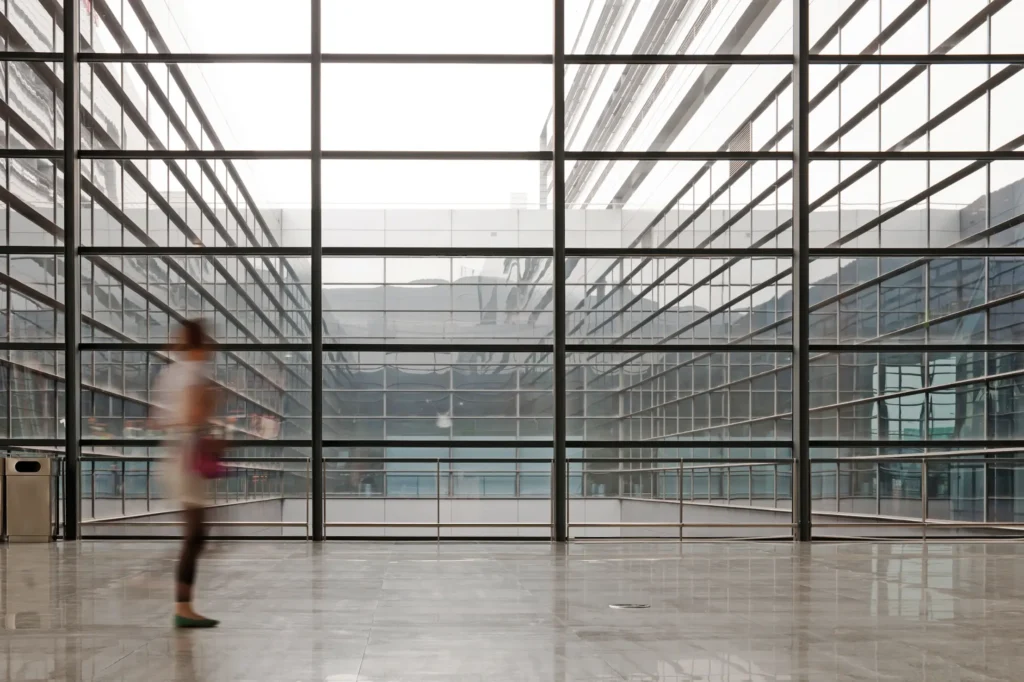 Blurred figure walking across a large open space with polished floors and expansive glass windows overlooking a modern building facade.