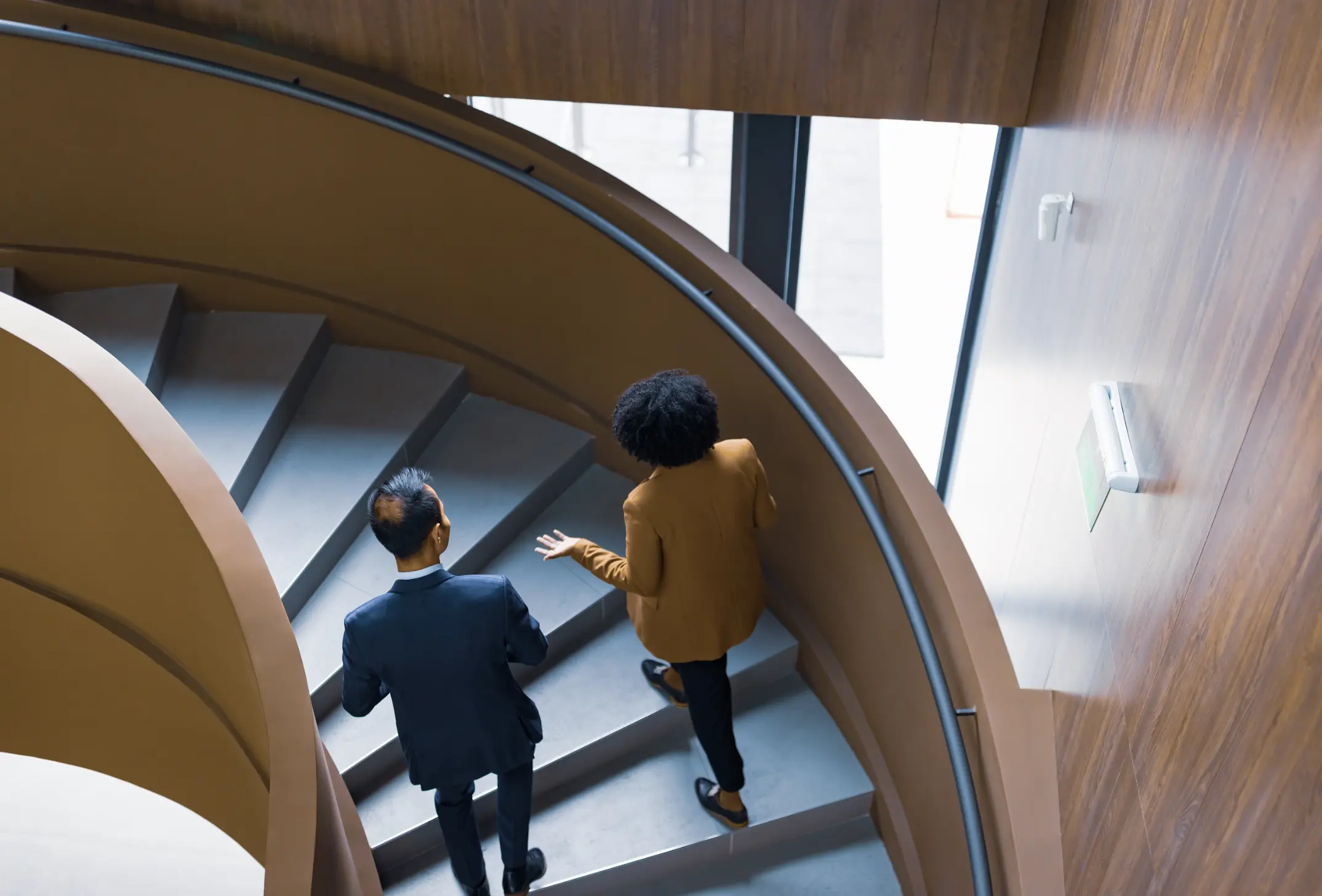Colleagues discussing ideas while ascending a spiral staircase in a contemporary office building with natural light.