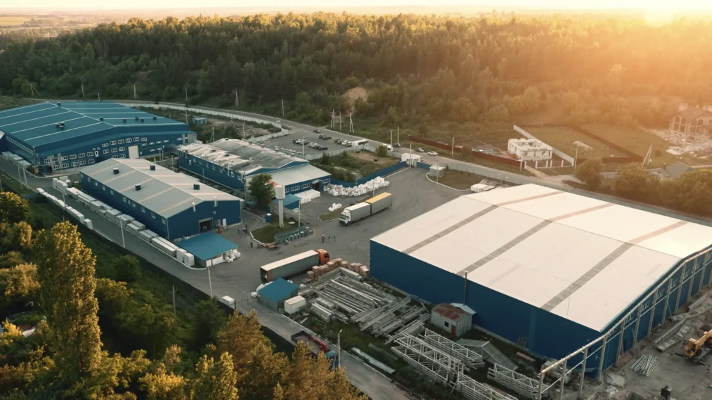 Aerial view of an industrial complex with large warehouses, shipping trucks, and surrounding forested area at sunset.
